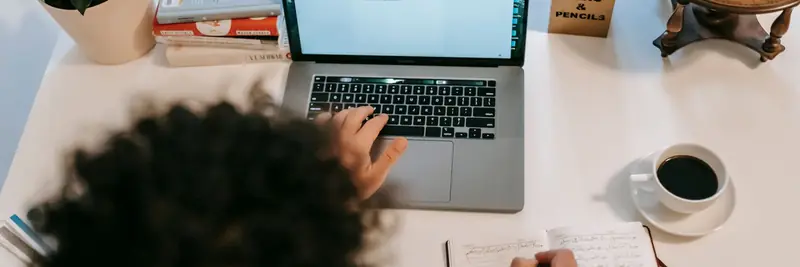 A writer typing on a laptop beside a notebook and a cup of coffee